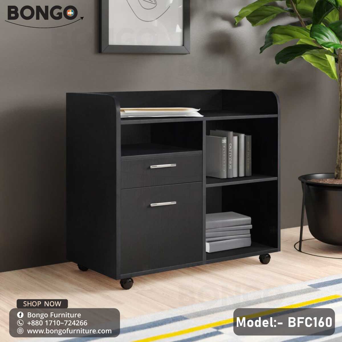A Black Enigma Drawer Unit with model number BFC160, featuring a single drawer and a lower shelf, set against a background of a living room with a potted plant and decorative items.