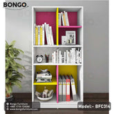 A modern white bookshelf from BONGO, featuring a playful, color-blocked design with bright magenta and yellow back panels and a central divider. The bookcase has an asymmetrical layout of open shelves, which are staged with books, binders, and decorative objects.
