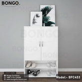 A white footwear shoe rack with multiple compartments, placed against a grey wall with a black shelf on top of it, displaying various shoes and a decorative plant.