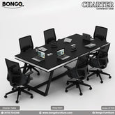A rectangular black conference table with a steel base, surrounded by black office chairs with wheels. The table has a white rectangular name plate on each side.