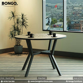 A round dining table with a beige laminated surface and a dark steel base, placed in a room with a view of buildings through the window.