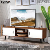 A promotional image of a modern TV stand in a well-lit living room setting. The low-profile console has a dark, reddish-brown wood-grain frame and top, contrasted with two solid white cabinet doors on either end. The center of the unit features two open shelves housing electronic devices. A large flat-screen television is mounted on the wall above the stand, displaying a scenic landscape. The room is styled with a tall, slender air conditioner to the right, a large potted plant to the left, and decorative i