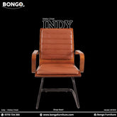 A promotional image of the 'INDY Visitor Chair,' a mid-century modern design. The chair features a single-piece bentwood shell in a dark wood finish, which includes integrated, curved armrests. The seat and back have tan, leather-style cushions with horizontal channel stitching, and the chair is supported by a black metal base. The image is set against a solid black background.