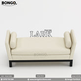 A promotional image of the 'LARK Divan Sofa', a modern, backless settee. The piece is upholstered in a cream-colored fabric and features high, flared arms with matching cylindrical bolster pillows. It is supported by a simple, recessed black frame and legs.