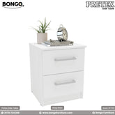 A modern white two-drawer nightstand shown in a bedroom setting. The simple, rectangular bedside table has two drawers with silver bar pulls and is decorated on top with a plant, books, and a classic silver alarm clock.