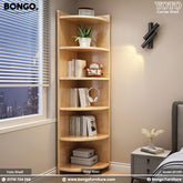 A white Yoto corner shelf with five tiers, featuring books, decorative items, and a potted plant on it, positioned in a room near a window with blinds.