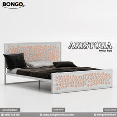 A promotional image of the "ARISTORA Metal Bed" by Bongo, shown with a white and rose-gold color scheme. The modern bed frame is made of white metal. The tall, rectangular headboard and matching lower footboard feature a decorative laser-cut pattern of organic, pebble-like shapes, which are backed by a light peachy-pink or rose-gold colored panel. The bed is made with contrasting dark brown and black bedding and is set against a plain white background.