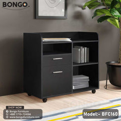 A Black Enigma Drawer Unit with model number BFC160, featuring a single drawer and a lower shelf, set against a background of a living room with a potted plant and decorative items.