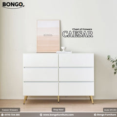 A promotional photograph of the "CAESAR Chest of Drawers" set in a bright, modern room. The wide, low dresser is finished in glossy white and features six handleless drawers arranged in two columns of three. It stands on four slender, angled, gold-colored legs. The dresser is styled with a leaning piece of minimalist art, a small stack of books, and is placed next to a potted plant and a sheer curtain. The "BONGO" brand logo is in the top-left corner.