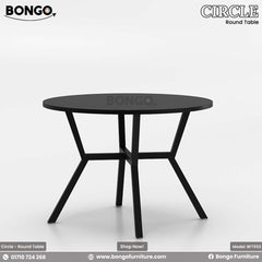 A round dining table with a beige laminated surface and a dark steel base, placed in a room with a view of buildings through the window.