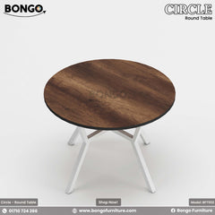 A round dining table with a beige laminated surface and a dark steel base, placed in a room with a view of buildings through the window.