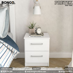 A modern white two-drawer nightstand shown in a bedroom setting. The simple, rectangular bedside table has two drawers with silver bar pulls and is decorated on top with a plant, books, and a classic silver alarm clock.