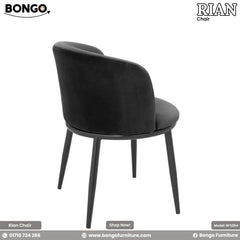 A close-up of a modern barrel chair, focusing on the curve of the back and arm. The chair is upholstered in a plush black velvet fabric, and the lighting highlights its soft texture and sheen. A piped seam runs along the top edge of the chair's curved form.