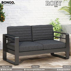 Gray sofa with black frame on a patio with plants and a white brick wall.
