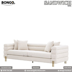 A modern three-seater sofa upholstered in a textured, off-white boucle fabric. The sofa's design is characterized by wide, horizontal channel tufting that wraps around its low back and arms. It is supported by slim, gold-finished metal legs and is shown with four matching throw pillows.