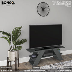 A promotional image of the "TEASER TV Stand" by Bongo, presented in a dark charcoal grey finish. The modern, geometric stand features two wide, angled A-frame style legs and a lower shelf. A large flat-screen television rests on top of the stand. The scene is set against a light grey wall, with a large, leafy potted plant on a stand to the left and a black, minimalist, sunburst-style clock hanging on the wall above. A patterned rug is visible on the floor.