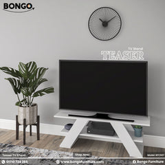 A promotional image of the "TEASER TV Stand" by Bongo, showcased in a modern living room setting. The TV stand is solid white and features a distinctive design with two wide, angled, A-frame style legs and a lower shelf. A large flat-screen television sits on top of the stand. To the left is a large potted plant in a grey pot on a dark wood stand, and on the light grey wall above is a black, minimalist, sunburst-style clock.